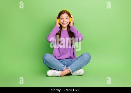 Full size photo of good mood girl with long hairdo dressed denim jeans palms touching headphones isolated on green color background Stock Photo