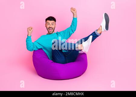 Full length photo of young nice guy sitting bean bag excited raise fists dressed stylish blue outfit isolated on pink color background Stock Photo