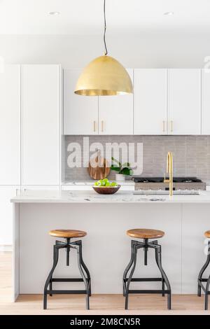 A kitchen detail with white cabinets, gold faucet and light hanging over the island with bar stools, and a tiled backsplash. Stock Photo