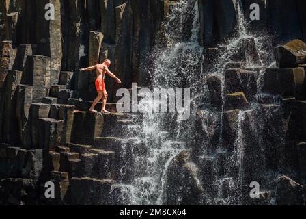 Caucasian man in swimsuit balancing near the falling water streams flowing on black volcanic stone cascades. Rochester Falls waterfall - popular touri Stock Photo
