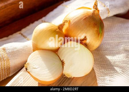 Unpeeled yellow onions with halved cut bulbs on wooden board background in the kitchen. Stock Photo