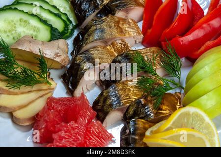 Slices of smoked fish mackerel or scomber on a white dish with lemon, apple, ginger, cucumber, bell pepper, grapefruit, dill, top view. Stock Photo