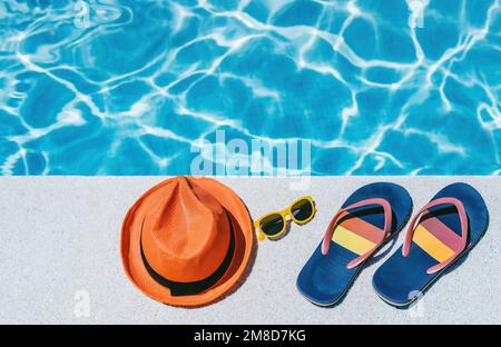 top view of yellow glasses, orange hat and flip-flops on the white edge of a swimming pool, with blue water in the background. concept of vacation Stock Photo