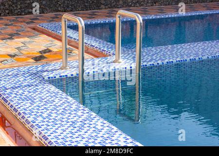 A metal swimming pool safe ladder (skimmer) on the entrance to the home inground poolside with the blue clean water. Stock Photo