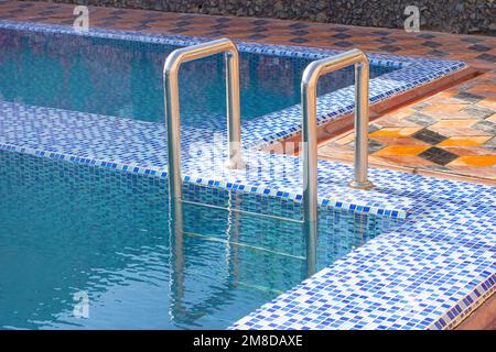 A metal swimming pool safe ladder (skimmer) on the entrance to the home inground poolside with the blue clean water. Stock Photo