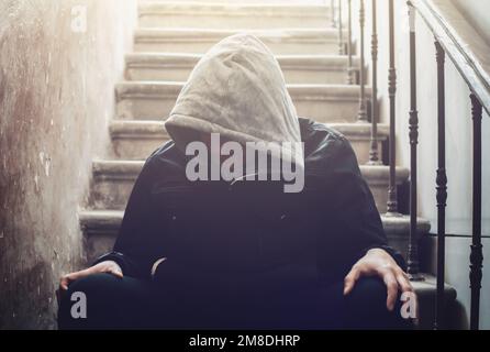 Lonely man in hood with his head down sits on stairs in dark. Stress and depression mental problems concept. Stock Photo