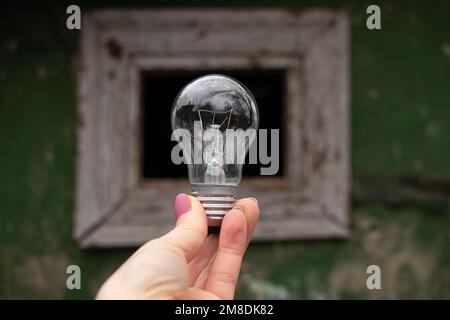 An incandescent light bulb in the hands of a girl against the background of an old window at home, people without light, light Stock Photo
