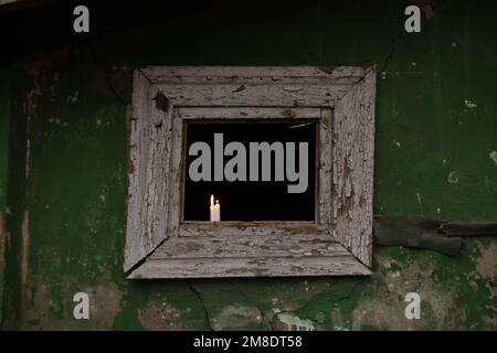 Three candles stand on a broken window of an old residential building, view from the street through the window, the light of a candle in a window in U Stock Photo
