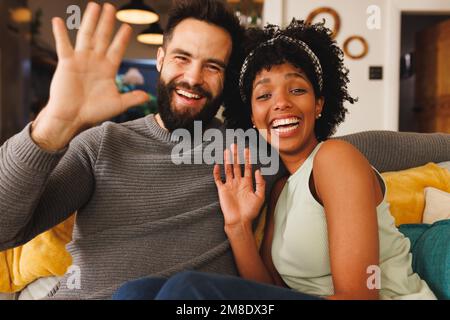 Portrait of cheerful biracial couple laughing and waving hands while relaxing on sofa in living room Stock Photo