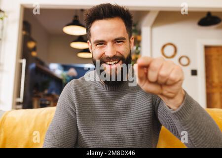 Portrait of biracial bearded young man pointing and laughing while sitting on sofa in living room Stock Photo