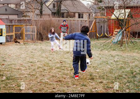 Two children play soccer with father in yard with chickens Stock Photo