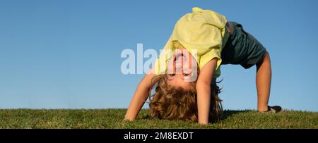 Happy little blond child upside down laying on the grass in the park. Smmer day during school holidays. Amazed kid boy dreaming and smiling. Active Stock Photo