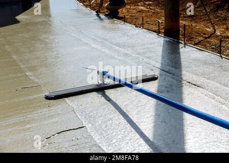 Worker is holding steel fresno trowel and smoothing out newly poured sidewalk on wet concrete in where construction taking place Stock Photo