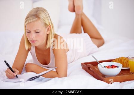 Shes got it all planned out...Beautiful woman lying on her bed and writing in her diary with her breakfast next to her. Stock Photo
