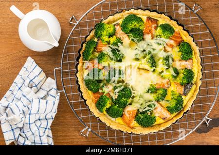 Top view of freshly baked delicious homemade broccoli and salmon quiche in black tart pan on metal cooling rack with white ceramic milk pot on wooden Stock Photo