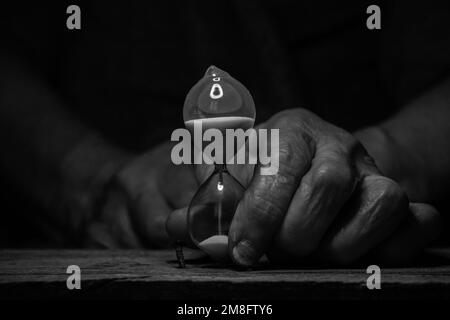 An old female hand holds an hourglass in the dark at the table, time goes by, the speed of life Stock Photo