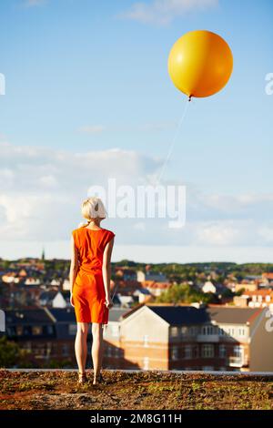 Celebrating her birthday with a balloon. Rearview of a gorgeous young woman in an orange dress with an orange balloon. Stock Photo
