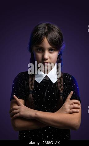 Portrait of little girl with Wednesday Addams costume during Halloween. Serious expression and dark atmosphere with dark background. Stock Photo