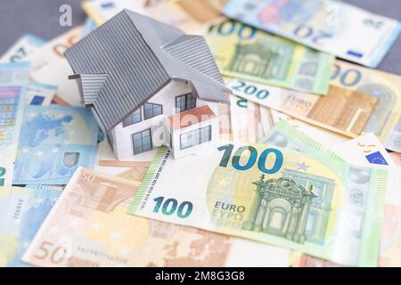 Small toy house is lies on a set of green monetary denominations of 100 euros. A lot of money forms an infinite heap. Stock Photo