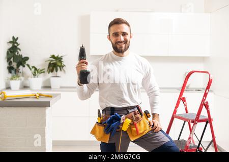 Home repairs. Portrait of professional male repairman with metal ladder. Smiling man in work overalls stands in middle of room in which he has Stock Photo