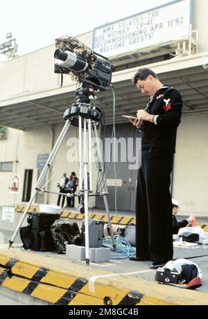 A Photographer's Mate 1ST Class (PH1) is captured on film making out the shoot sheets which are as necessary as exposing the film. In front of him is a video camera mounted on a tripod to capture the event. He is covering the decommissioning of the aircraft carrier USS Midway (CV-41) which served the nation for forty-seven years. Base: Naval Air Station, North Island State: California (CA) Country: United States Of America (USA) Stock Photo