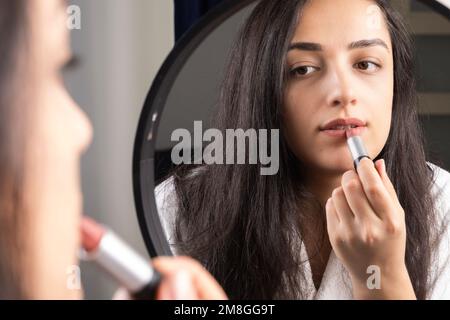 Beautiful millennial woman looking mirror and doing make up. Brunette caucasian girl rouging lipstick. Every day morning routine. Applying cosmetics. Stock Photo