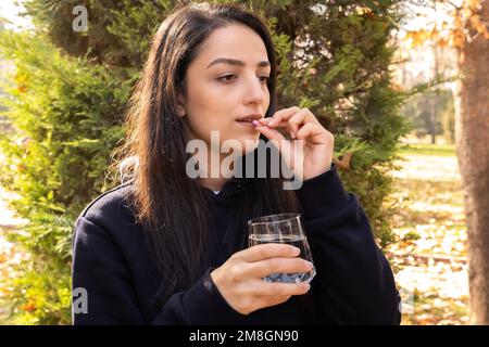 Unhappy woman holding glass of water and taking pills. Side view portrait of female taking painkiller for ache concept idea. Copy space. Upset face. Stock Photo