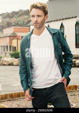 Young handsome guy is standing on a western town background. Man is wearing white empty t-shirt without logo. Vertical mock-up. Stock Photo