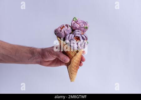 Mans hand holds a waffle cone with sweet pastel purple marshmallow peonies on a light isolated background. Mock up, copy space. Small business, pastry Stock Photo
