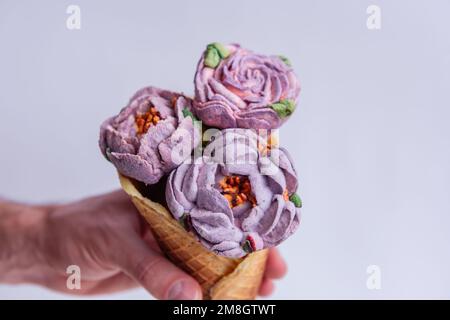 Mans hand holds a waffle cone with sweet pastel purple marshmallow peonies on a light isolated background. Mock up, copy space. Small business, pastry Stock Photo