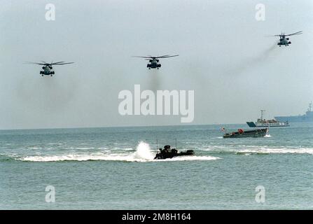 CH-53E Super Stallion helicopters, left, and a CH-53 Sea Stallion helicopter, right, fly overhead as an AAVP-7A1 amphibious assault vehicle, foreground, and a mechanized landing craft (LCM) assigned to the tank landing ship USS MANITOWOC (LST-1100) come ashore during an amphibious assault exercise. In the background, utility landing craft LCU-1653 brings Marines toward shore as an amphibious assault ship stands at anchor. Country: Unknown Stock Photo