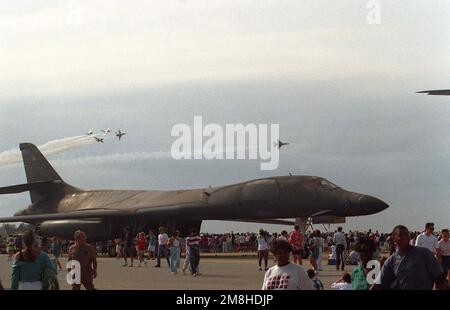 A right side view of a B-1B Lancer aircraft on display during an air show. In the background the Thunderbirds flight demonstration team performs an aerial maneuver for the crowd. Base: Eglin Air Force Base State: Florida (FL) Country: United States Of America (USA) Stock Photo