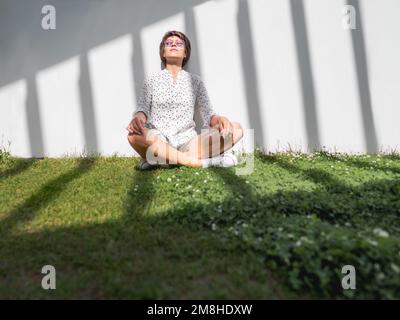 Woman is meditating on lawn in urban park. Nature in town. Relax outdoors after work. Summer vibes. Stock Photo