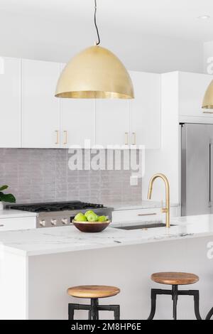 A kitchen detail with white cabinets, gold faucet and light hanging over the island with bar stools, and a tiled backsplash. Stock Photo