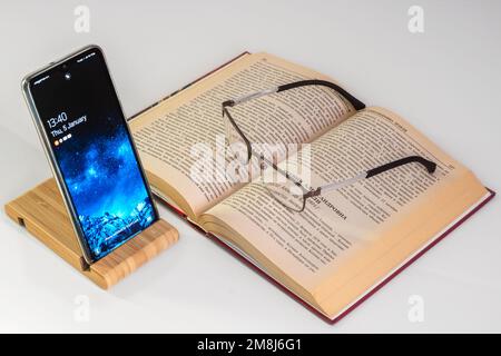 Switched on mobile phone installed on a special stand and an open book with glasses on a white background. Stock Photo
