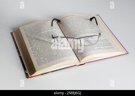 Open book with glasses on top of the pages lies on a white table surface. Stock Photo
