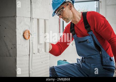 Caucasian Construction Contractor Worker in his 40s Checking on Newly Installed Brick Wall Power Outlet Stock Photo
