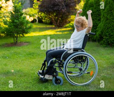 Elderly woman stretches her arms up while sitting in a wheelchair on a walk outdoors.  Stock Photo