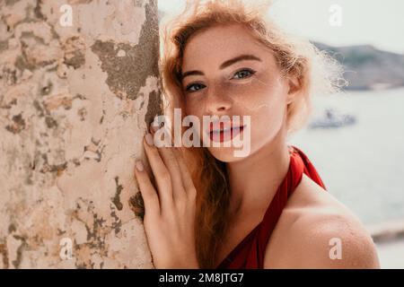 Redhead woman portrait. Curly redhead young caucasian woman with freckles looking at camera and smiling. Cute woman close up portrait in a red long Stock Photo