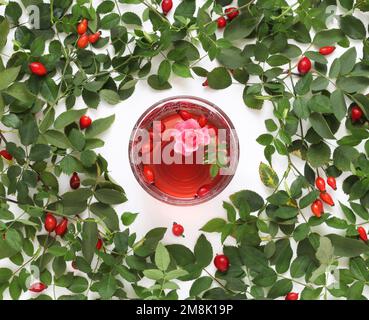 Cup of red rosehip tea on background with fresh leaves and berries, table top view. Glass mug of herbal healthy tea with rosehips, leaves and flower. Stock Photo