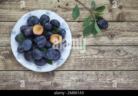 Fresh blue plums in bowl on wooden table background with leaves and twig, halved plum fruit with seed as decoration, top view. Stock Photo