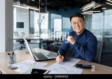 A successful young man businessman Asian works in the office at the table, works with documents, graphs, plan, accounts. Writes, looks at the camera, smiles. Stock Photo