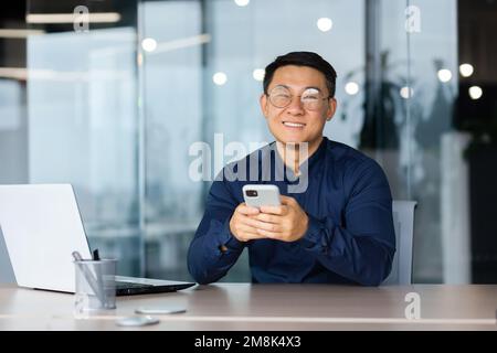 A young man in glasses sits in the office at a table with a laptop, holds a phone in his hands, smiles at the camera. Stock Photo