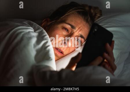 Young woman uses smartphone while lying in bed at night. Insomnia and social media addiction concept. Stock Photo