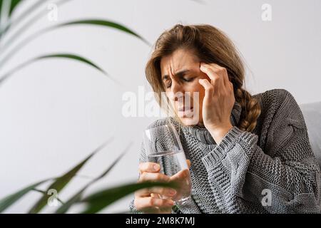 Headache. Young woman, who is sitting on a sofa with her eyes closed, touching her head while suffering from a migraine and holding a glass of water. Stock Photo