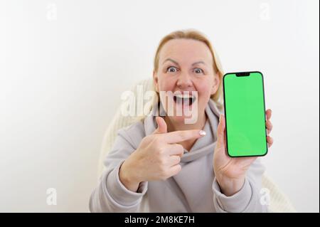 pretty sweet woman in white hoodie looks at the phone, she is glad that she sees there and shows the phone with green screen to camera. Happy lady sha Stock Photo