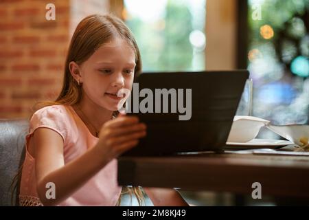 Smiling preteen girl sitting at restaurant table and watching video of her favorite blogger on tablet computer Stock Photo