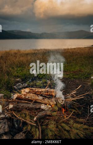 A small campfire with gentle flames beside a lake during a glowing sunset Stock Photo