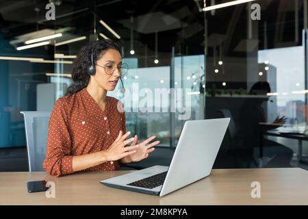 Video call online meeting with colleagues, Hispanic woman working inside modern office, businesswoman smiling and talking remotely using laptop and headset. Stock Photo