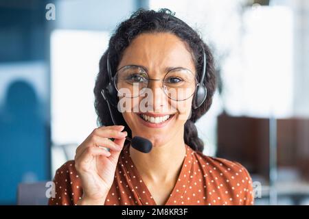 Close up portrait of latin american woman inside modern office with headset for video call, woman smiling and looking at camera, office worker customer support tech helpline. Stock Photo